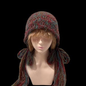 Puede incluir: Un gorro tejido a mano, multicolor, con orejeras y largas borlas. El gorro presenta una mezcla de hilos rojos, turquesa y marrones. Las borlas cuelgan a ambos lados de la cara. Se muestra sobre una cabeza de maniquí sobre un fondo negro.