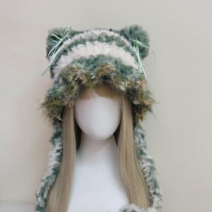 May include: A whimsical, furry hat in shades of green, white, and brown, designed with cat ears and ribbons. The hat has a striped pattern and a fluffy texture, creating a playful and unique accessory.