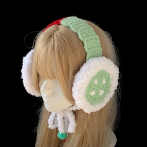 May include: Festive crocheted earmuffs. The headband is green with a red stripe, and the ear coverings are white with a green design. A white, fluffy scarf with a green and red pom-pom hangs below the earmuffs.