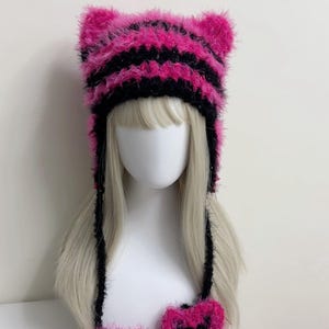 K&ouml;nnte beinhalten: Ein rosa-schwarz gestreifter Katzenohrhut mit passenden fingerlosen Handschuhen. Der Hut und die Handschuhe sind aus einem flauschigen, strukturierten Material gefertigt. Der Hut hat zwei aufrechte Katzenohren und lange schwarze B&auml;nder.