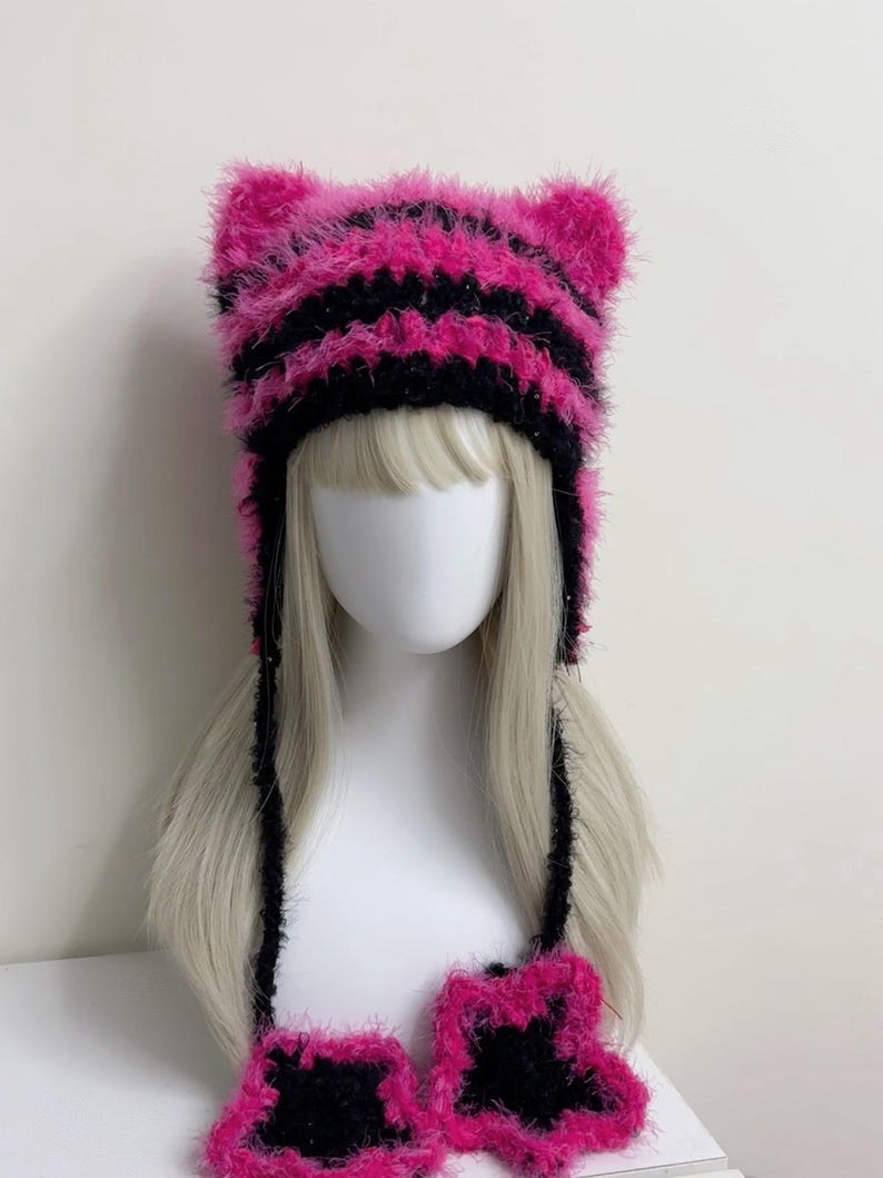 K&ouml;nnte beinhalten: Ein rosa-schwarz gestreifter Katzenohrhut mit passenden fingerlosen Handschuhen. Der Hut und die Handschuhe sind aus einem flauschigen, strukturierten Material gefertigt. Der Hut hat zwei aufrechte Katzenohren und lange schwarze B&auml;nder.