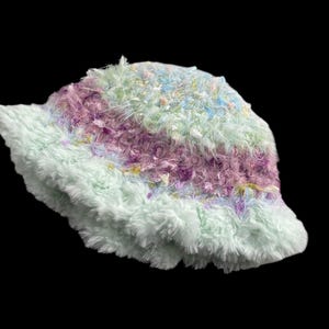May include: A fluffy bucket hat in pastel colors. The hat features a soft, textured material in shades of light green, purple, and blue. The brim and body of the hat are made of a plush, fuzzy fabric.