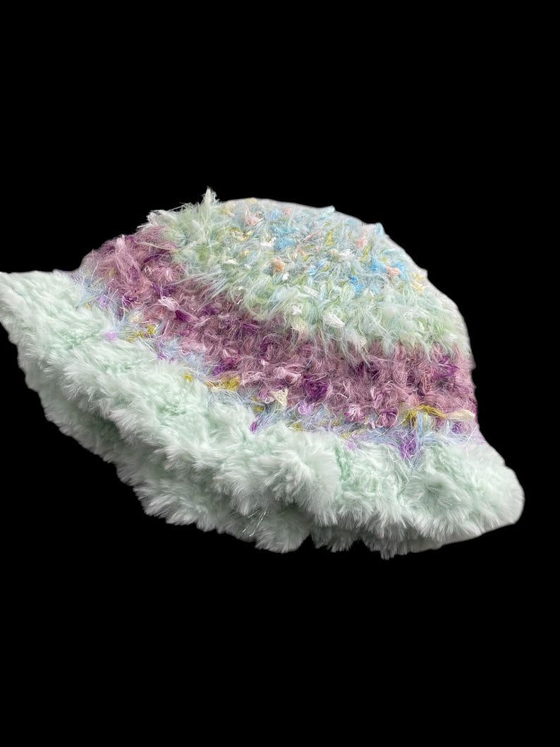 May include: A fluffy bucket hat in pastel colors. The hat features a soft, textured material in shades of light green, purple, and blue. The brim and body of the hat are made of a plush, fuzzy fabric.