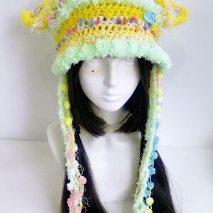 May include: A whimsical, handmade crochet hat in shades of yellow, green, and pastel colors. The hat features a brim, ear flaps, and long, decorative strands with pom-poms. A blue heart-shaped button adorns the front. The hat is displayed on a mannequin head.
