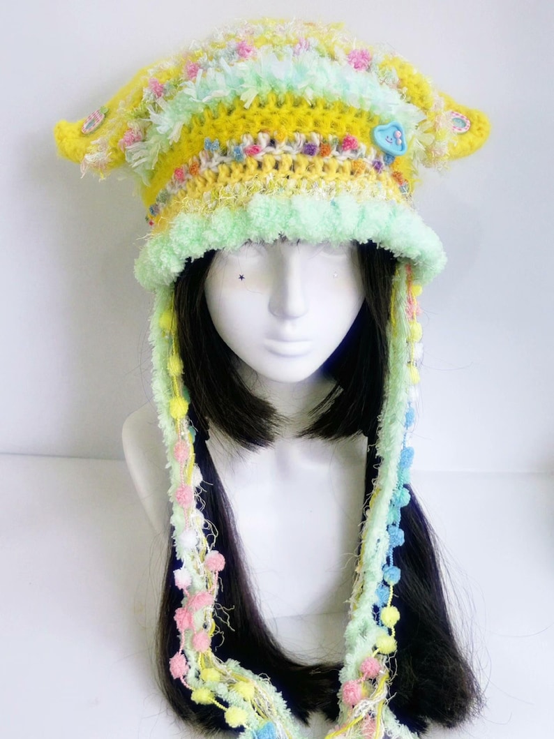 May include: A whimsical, handmade crochet hat in shades of yellow, green, and pastel colors. The hat features a brim, ear flaps, and long, decorative strands with pom-poms. A blue heart-shaped button adorns the front. The hat is displayed on a mannequin head.