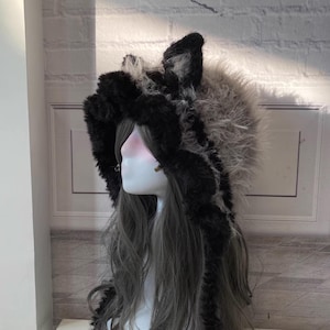 May include: A black and gray faux fur hooded hat with long, dangling fur tails. The hat has two furry ears on top.