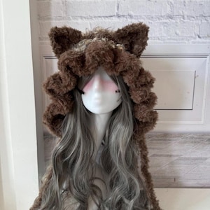 May include: A brown faux fur hood with attached ears and a ruffled trim. The hood is worn over a wig of long, wavy gray hair. The mannequin head has a painted face with pink blush and black accents.