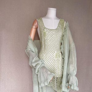 Puede incluir: Un vestido de crochet verde claro con un cárdigan a juego de manga larga. El vestido presenta un patrón de red y detalles de flecos. El cárdigan tiene mangas con volantes. El conjunto se exhibe en un maniquí.