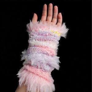 May include: A colorful, handmade wrist warmer in shades of pink, purple, and white. The knitted design features a fluffy, feathery trim at the top and bottom. The wrist warmer is a unique accessory.