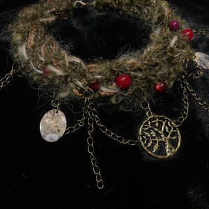 May include: A handcrafted necklace featuring a woven, earthy-toned band adorned with red and green beads. Antique gold-colored chain dangles with oval and tree-shaped pendants. The necklace is designed for a bohemian or nature-inspired aesthetic.