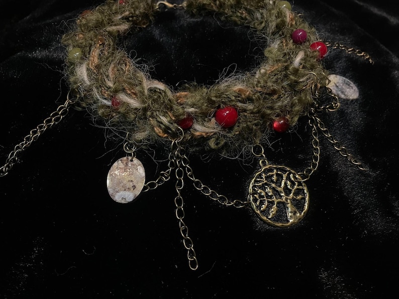May include: A handcrafted necklace featuring a woven, earthy-toned band adorned with red and green beads. Antique gold-colored chain dangles with oval and tree-shaped pendants. The necklace is designed for a bohemian or nature-inspired aesthetic.