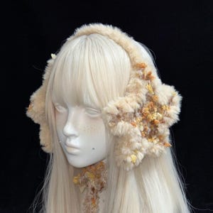 Pode incluir: Uma faixa de orelhas de pele sintética bege com detalhes florais amarelos e laranja. As orelhas são exibidas em uma cabeça de manequim com cabelo loiro longo e liso em um fundo preto. Um acessório de inverno.