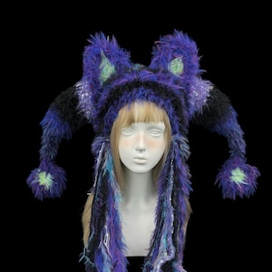 Pode incluir: Um chapéu extravagante, feito à mão, em tons de roxo, preto e azul, com longas mechas penduradas e protuberâncias em forma de orelhas. O chapéu tem uma textura fofa e é adornado com pequenos detalhes verdes. O chapéu é exibido em uma cabeça de manequim sobre um fundo preto.