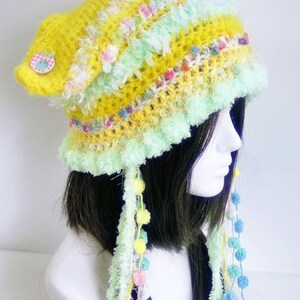 May include: A handmade, whimsical crochet hat in shades of yellow, green, and pastel colors. The hat features a button detail, fluffy trim, and dangling yarn with pom-poms. This unique accessory adds a playful touch to any outfit.