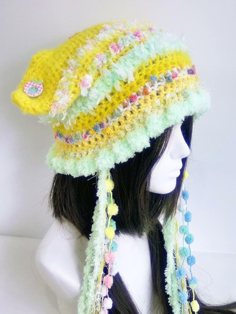 May include: A handmade, whimsical crochet hat in shades of yellow, green, and pastel colors. The hat features a button detail, fluffy trim, and dangling yarn with pom-poms. This unique accessory adds a playful touch to any outfit.