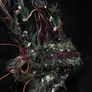 May include: Close-up of a handcrafted, textured floral arrangement. The piece features a mix of green, burgundy, and white elements, with brown leather accents. The arrangement is set against a black background, highlighting the intricate details.