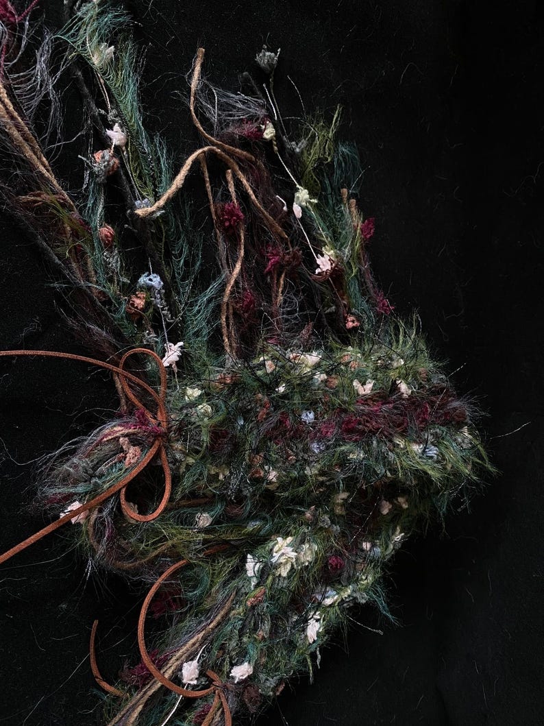 May include: Close-up of a handcrafted, textured floral arrangement. The piece features a mix of green, burgundy, and white elements, with brown leather accents. The arrangement is set against a black background, highlighting the intricate details.