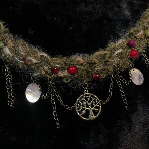 May include: A handcrafted necklace featuring a thick, textured, olive-green yarn base adorned with red and green beads. Silver-toned chains dangle from the base, suspending oval pendants and a tree of life charm. The necklace is designed for a unique, bohemian style.