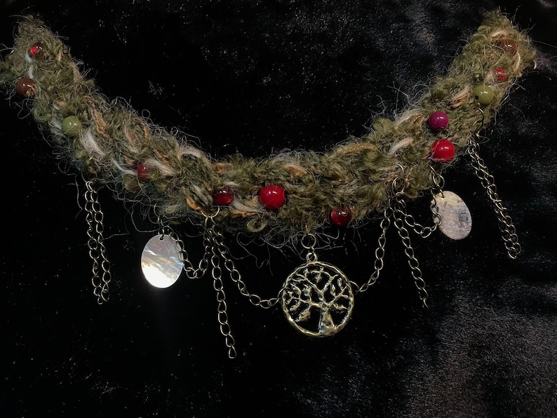 May include: A handcrafted necklace featuring a thick, textured, olive-green yarn base adorned with red and green beads. Silver-toned chains dangle from the base, suspending oval pendants and a tree of life charm. The necklace is designed for a unique, bohemian style.
