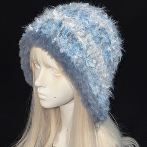 May include: A soft, fluffy hat in shades of light blue, with a darker blue brim. The hat has a textured appearance, with white and blue fibres woven throughout. This winter hat is designed to be worn on the head.
