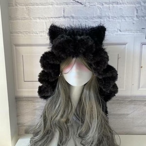 Puede incluir: Un gorro negro de orejas de gato de piel sintética con pelo largo y ondulado de color gris. El gorro presenta orejas erguidas y una textura esponjosa. La peluca está peinada sobre una cabeza de maniquí.