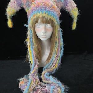 May include: A colourful jester hat with long, flowing strands. The hat features a mix of pastel colours, including pink, blue, yellow, and purple. The hat is made of a fuzzy, textured material and has two pointed horns.