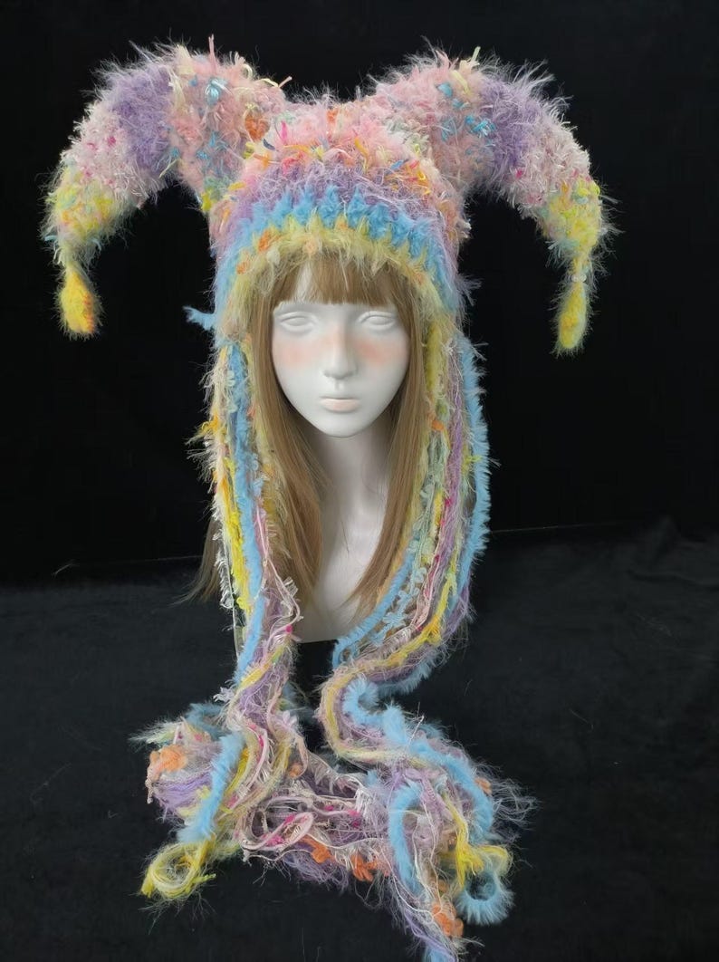 May include: A colourful jester hat with long, flowing strands. The hat features a mix of pastel colours, including pink, blue, yellow, and purple. The hat is made of a fuzzy, textured material and has two pointed horns.