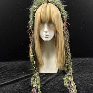 May include: A green and brown headpiece with long, decorative strands. The headpiece is adorned with small flowers and textured materials. The headpiece is displayed on a mannequin head with long, blonde hair.