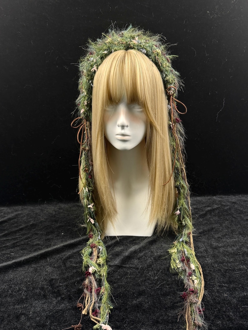 May include: A green and brown headpiece with long, decorative strands. The headpiece is adorned with small flowers and textured materials. The headpiece is displayed on a mannequin head with long, blonde hair.