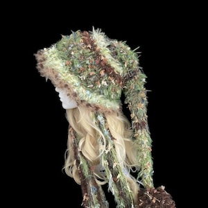 May include: A long, pointed hat crafted from textured, shaggy yarn in shades of forest green, brown, and cream. The hat features a wide, fuzzy brim and long, dangling side panels that terminate in large, dense pom-poms of the same mixed-fiber yarn.