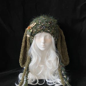 May include: A whimsical green hat with long, floppy ears, adorned with floral accents and a small bunny figure. The hat is displayed on a mannequin head with white hair, set against a dark background. The hat's texture appears soft and fuzzy.