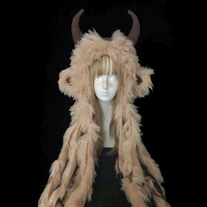 Puede incluir: Una capucha de disfraz de piel sintética beige con cuernos marrones y pelo largo y fluido. La capucha está diseñada para parecerse a una criatura mítica. El disfraz se exhibe en un maniquí con pelo largo y rubio, sobre un fondo negro.