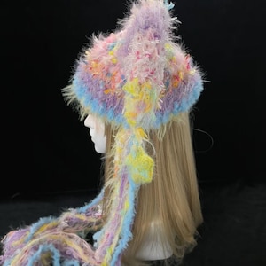May include: A handmade hat with ear flaps and long strands, in pastel shades. The fluffy, textured design incorporates lavender, blue, yellow, and pink. The hat is designed to be worn, and is a unique fashion accessory.