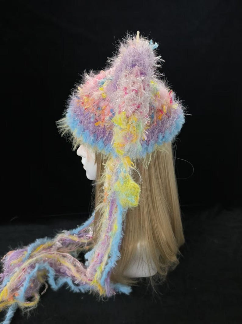 May include: A handmade hat with ear flaps and long strands, in pastel shades. The fluffy, textured design incorporates lavender, blue, yellow, and pink. The hat is designed to be worn, and is a unique fashion accessory.