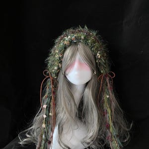 May include: A wig with long, gray hair and a decorative headpiece. The headpiece is made of green and brown materials with small flowers and accents. Brown ribbons and strands of the headpiece hang down the sides.