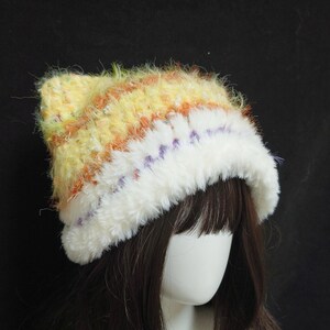 May include: A handmade, fluffy hat with a white brim and a yellow, orange, and purple striped crown. The hat is made of soft, textured yarn, perfect for cold weather. The hat is shown on a mannequin head with dark brown hair.