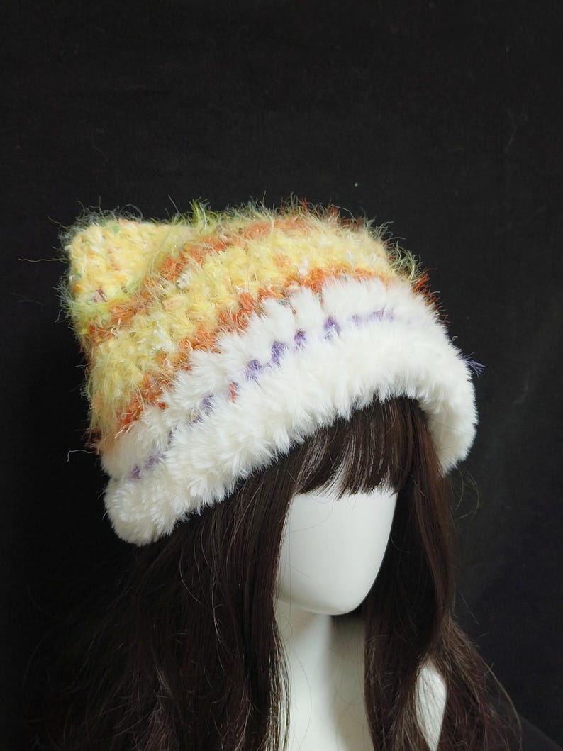 May include: A handmade, fluffy hat with a white brim and a yellow, orange, and purple striped crown. The hat is made of soft, textured yarn, perfect for cold weather. The hat is shown on a mannequin head with dark brown hair.