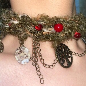 May include: A handmade choker necklace with a textured, olive green base. The necklace features red beads, dangling chains, and decorative pendants, including a tree of life design. The metal accents have a bronze finish.