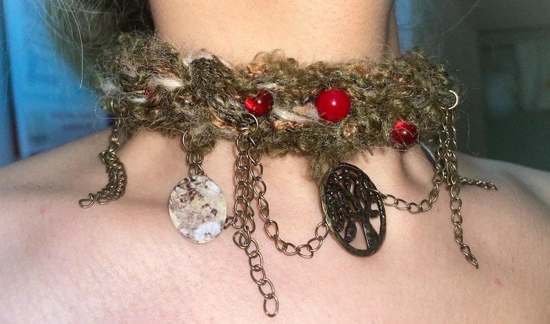 May include: A handmade choker necklace with a textured, olive green base. The necklace features red beads, dangling chains, and decorative pendants, including a tree of life design. The metal accents have a bronze finish.