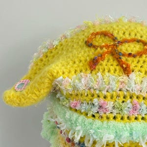 May include: A handmade, yellow crocheted hat with an orange bow and decorative trim. The hat features a small, pink and white gingham patch on the brim. The trim includes pastel colors like pink, green, and white. The hat is a unique, handcrafted item.