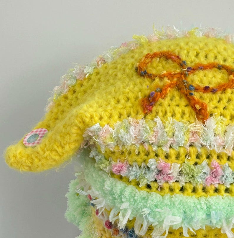 May include: A handmade, yellow crocheted hat with an orange bow and decorative trim. The hat features a small, pink and white gingham patch on the brim. The trim includes pastel colors like pink, green, and white. The hat is a unique, handcrafted item.