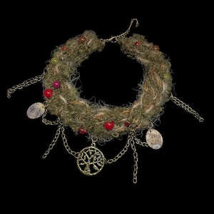 May include: A handcrafted necklace with a woven, moss-green base adorned with red beads and dangling chains. The chains hold small, oval pendants and a tree-of-life charm, all against a black background.