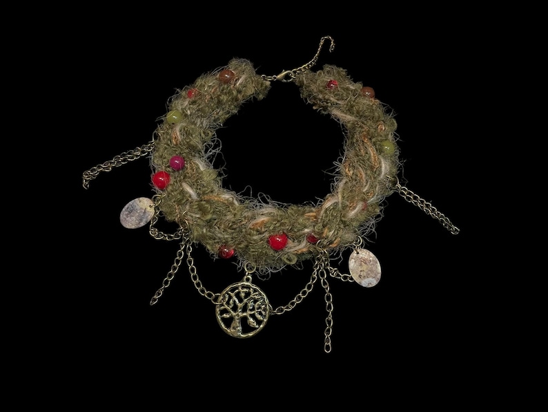 May include: A handcrafted necklace with a woven, moss-green base adorned with red beads and dangling chains. The chains hold small, oval pendants and a tree-of-life charm, all against a black background.