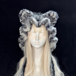 May include: A fluffy, faux fur hat featuring a mottled gray, black, and white pattern with two prominent, rounded ear-like projections on the top. The hat has a thick, textured pile and frames the face, showcasing a soft, plush appearance.