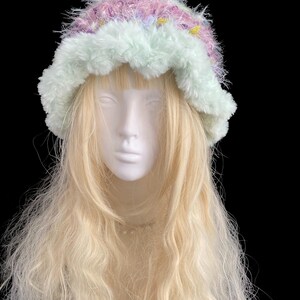 May include: A fluffy, pastel-colored hat with a light green brim and a crown of pink, purple, and yellow. The hat is made of a soft, textured material and is displayed on a mannequin head with long, blonde hair.