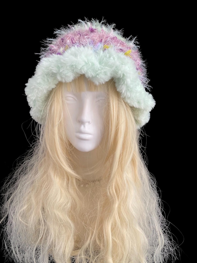 May include: A fluffy, pastel-colored hat with a light green brim and a crown of pink, purple, and yellow. The hat is made of a soft, textured material and is displayed on a mannequin head with long, blonde hair.
