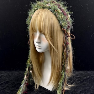 May include: A green and burgundy floral crown with long, flowing strands. The crown is made of various textures and colors, including green, burgundy, and cream-colored accents. The crown is displayed on a mannequin head with blonde hair.