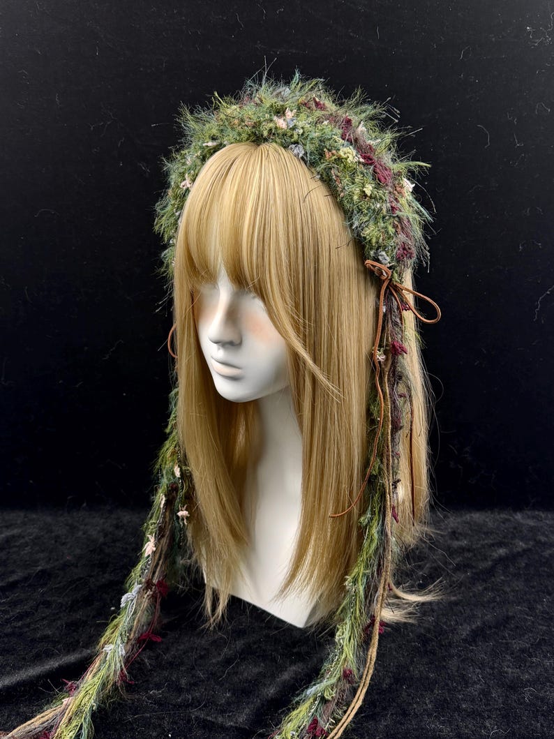 May include: A green and burgundy floral crown with long, flowing strands. The crown is made of various textures and colors, including green, burgundy, and cream-colored accents. The crown is displayed on a mannequin head with blonde hair.