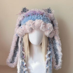 May include: A plush, fuzzy hat featuring long ear-like flaps, rendered in a soft palette of pastel pink, light blue, and white. The hat includes decorative pink ribbons tied at the top and textured, multi-colored yarn accents hanging down.