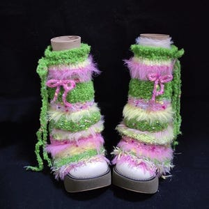May include: A pair of platform boots with colorful leg warmers. The leg warmers are green, pink, and yellow, with a fluffy texture and a pink bow. The boots have white uppers and a brown sole.